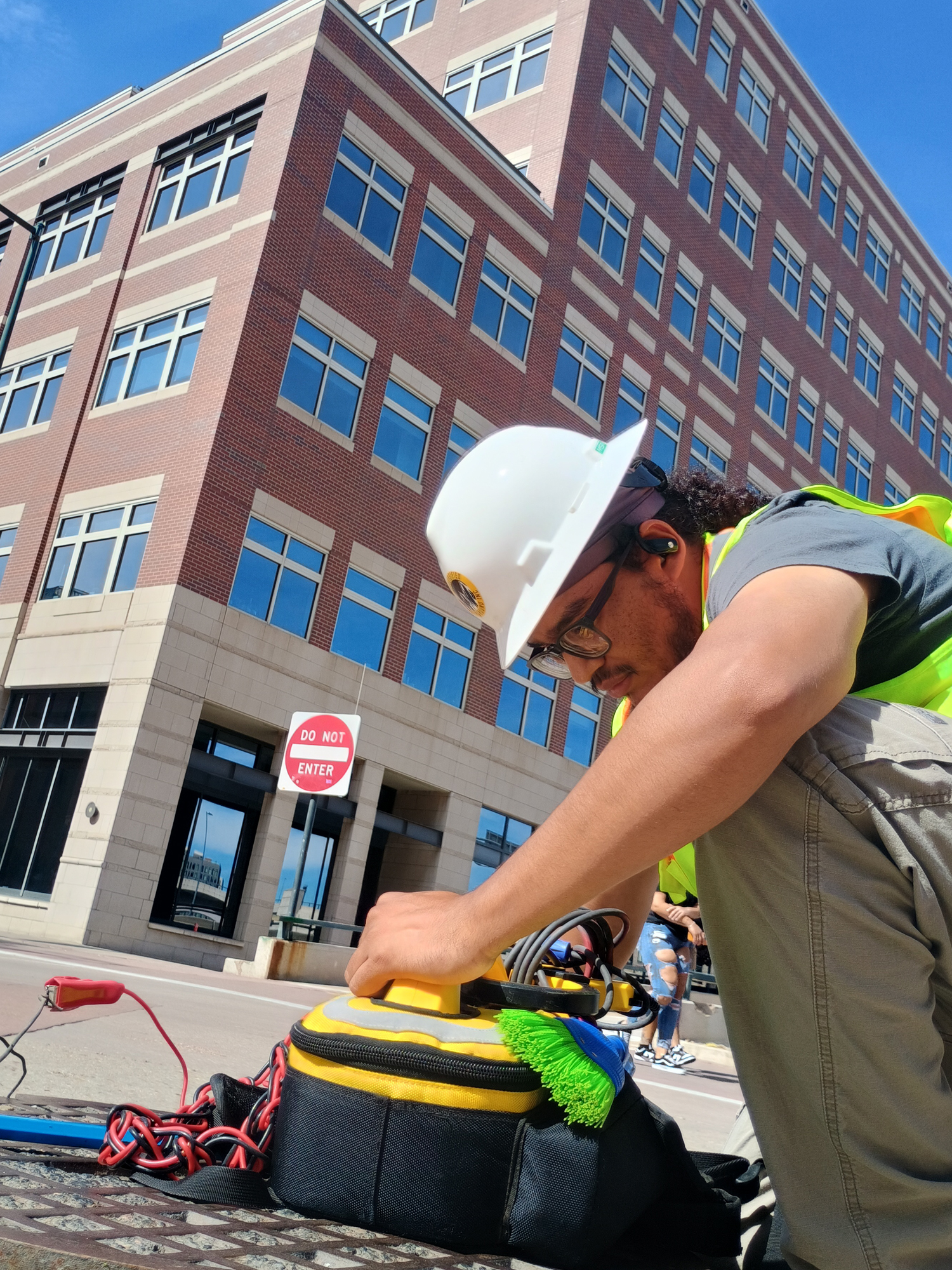 du-crew-close-up-urban A construction worker in a hard hat and safety vest uses equipment on a city sidewalk near a red brick building, with a Do Not Enter sign visible in the background.