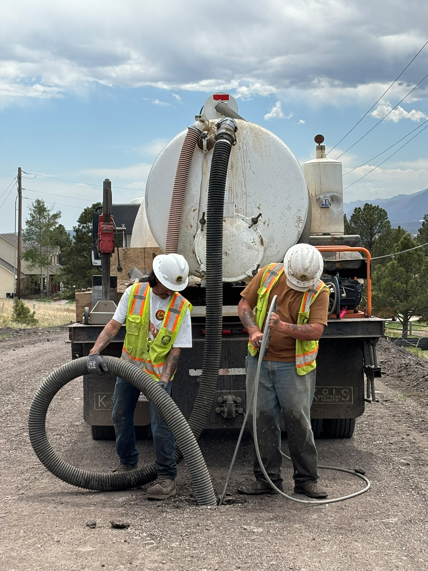 du-crew-wide-hose Two construction workers in safety vests and helmets operate hoses connected to a large tank truck, performing road maintenance on a dirt street under a cloudy sky.