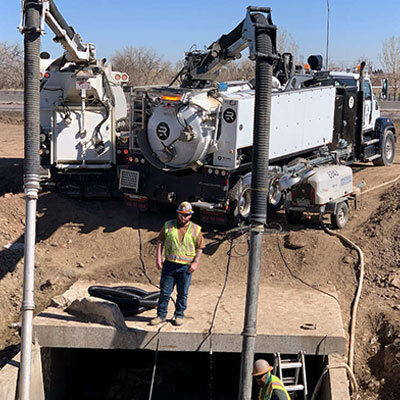 Two workers stand near large vacuum trucks at a construction site, working around a concrete drainage culvert. One worker is on the culvert, and the other is inside. Equipment pipes extend into the culvert.