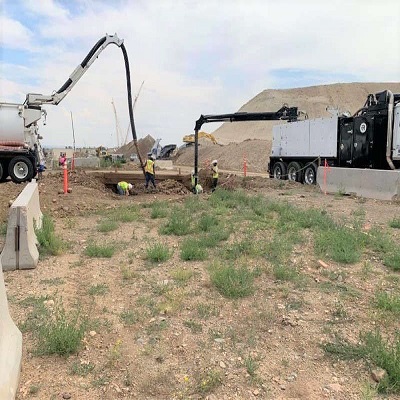 Three construction workers use heavy machinery to dig and move dirt in a dry, open area with trucks and mounds of earth in the background under a partly cloudy sky.