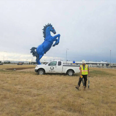 A worker in a safety vest walks toward a white pickup truck parked near a large blue rearing horse statue on a grassy area, with buildings and a cloudy sky in the background.