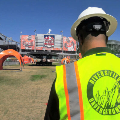 A worker in a hard hat and safety vest labeled Diversified Underground stands on a grassy field near a stadium with orange tents and a large sign featuring football players in the background.
