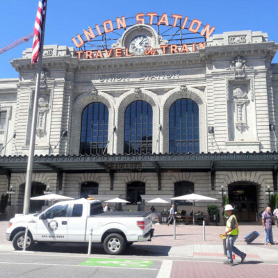 The front of Union Station with large arched windows, a pickup truck, a person in a safety vest, American flag, and outdoor seating with umbrellas on a sunny day.