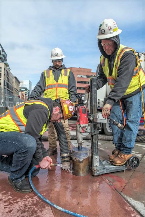Three construction workers in safety gear use a drill to core into concrete on a city street. One operates the drill while the others assist, with buildings and equipment in the background.