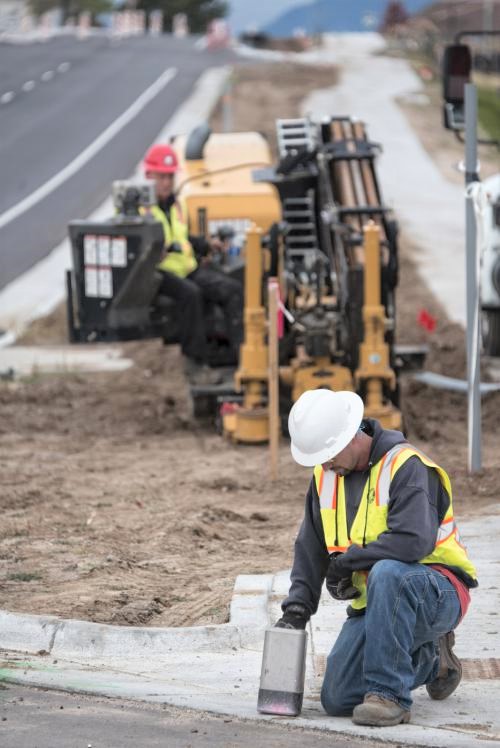 Two construction workers in safety gear work near a road; one operates machinery in the background while the other kneels on the sidewalk, using a tool on the concrete.