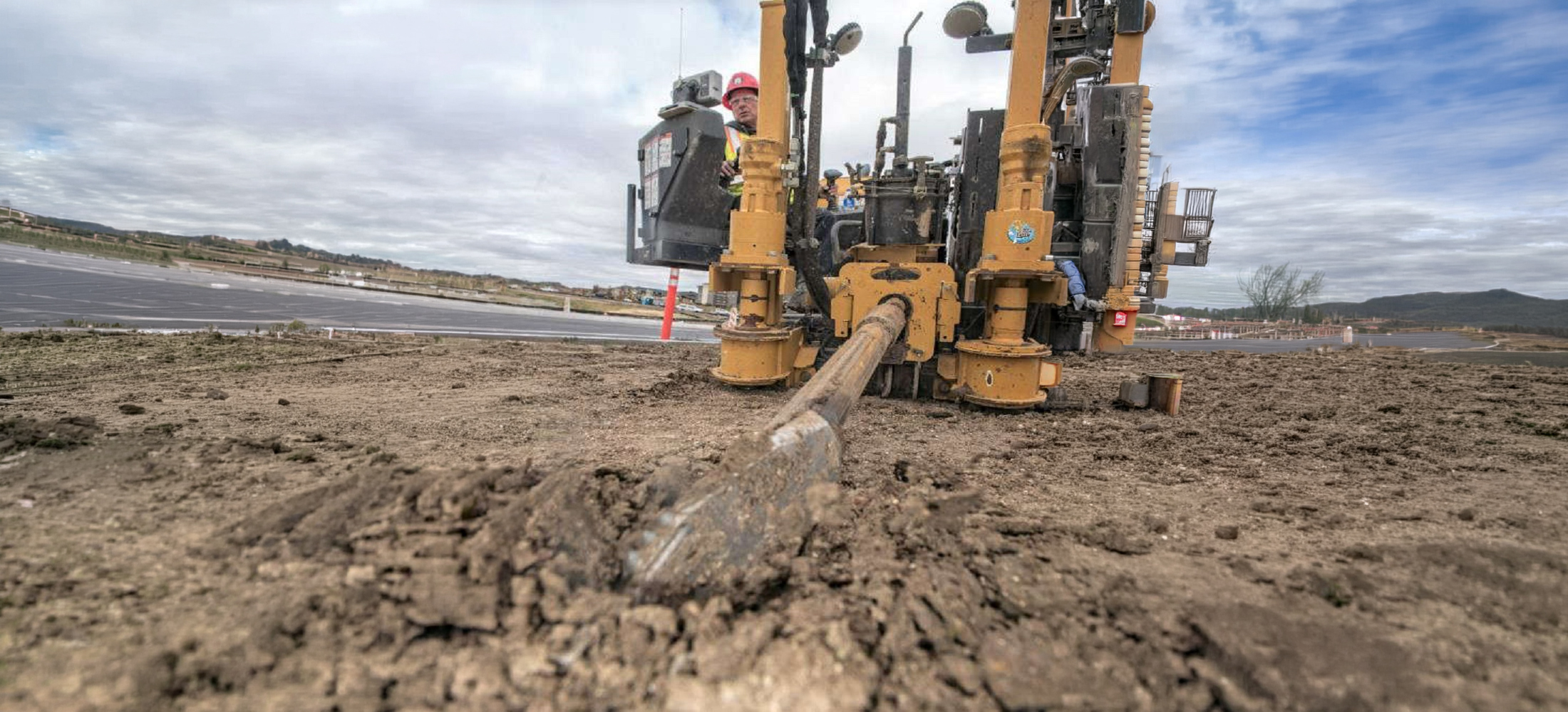 A worker operates a large yellow drilling machine on a construction site, drilling into the dirt under a cloudy sky.