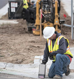 A construction worker in a white hard hat and safety vest kneels on a curb, using a tool. Construction equipment and another worker are visible in the background.