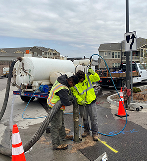 Two workers in safety gear use equipment to clean or inspect a utility hole on a street. A white utility truck and traffic cones are nearby, and houses and cloudy sky are visible in the background.