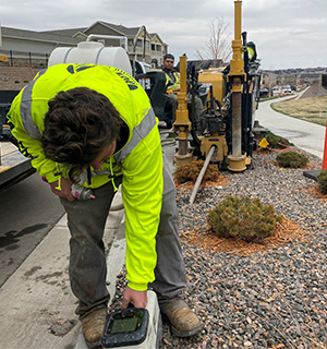 A worker in a neon yellow jacket checks a device on a curb near landscaping rocks, with construction equipment and another worker visible in the background on a cloudy day.