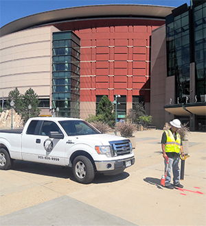 A worker in a yellow safety vest uses equipment near a white pickup truck parked outside a large modern building with red and glass exterior walls.