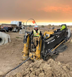 A construction worker in a safety vest operates a Vermeer drilling machine at a dusty worksite during sunset, with trucks and concrete pipes in the background.