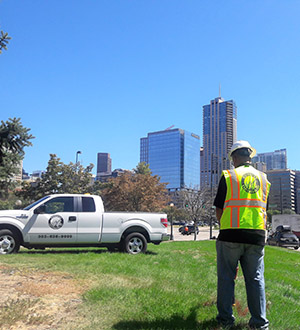 A worker in a safety vest and hard hat stands on grass near a white pickup truck, facing a city skyline with tall buildings under a clear blue sky.