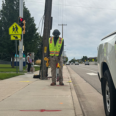 A construction worker in a safety vest and helmet stands on a sidewalk near a road, holding tools. A white truck is parked nearby, and a pedestrian crossing sign is visible in the background.