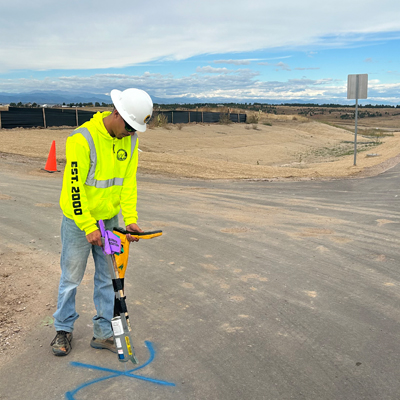 A construction worker in a neon yellow hoodie and hard hat uses a detection tool on a paved road marked with blue paint, with dirt, fencing, and a traffic cone in the background.