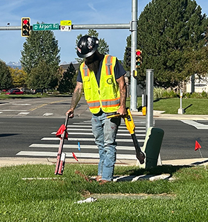 A construction worker in a safety vest uses equipment to locate underground utilities near a street intersection, with red flags marking the area and traffic lights visible in the background.