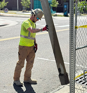 A construction worker wearing a hard hat, sunglasses, and safety vest steadies a large concrete post next to a fenced area on a sunny day.