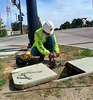 A construction worker in a safety vest and hard hat repairs electrical wires at an open utility box near a street intersection on a sunny day.