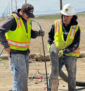 Two construction workers in yellow safety vests and helmets work outdoors with hoses and cables on a dirt site, with fields and utility poles visible in the background.