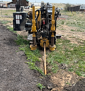 A yellow horizontal directional drilling machine is operating outdoors, boring a hole into the ground with a metal rod, surrounded by grass, dirt, and a few buildings in the background.